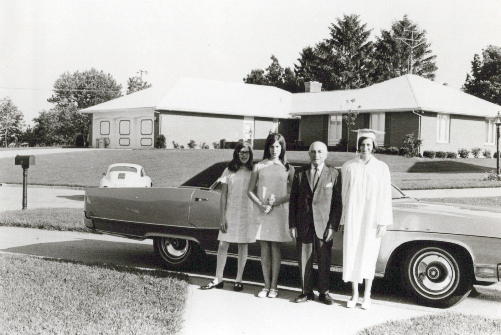 A black-and-white photograph of a graduate in cap and gown beside her family and a long sedan, a ranch house behind them