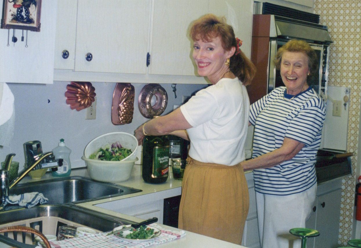 A colour snapshot of two women cooking in a kitchen: one tosses a salad by the sink and smiles back at the camera, an older woman laughs behind her