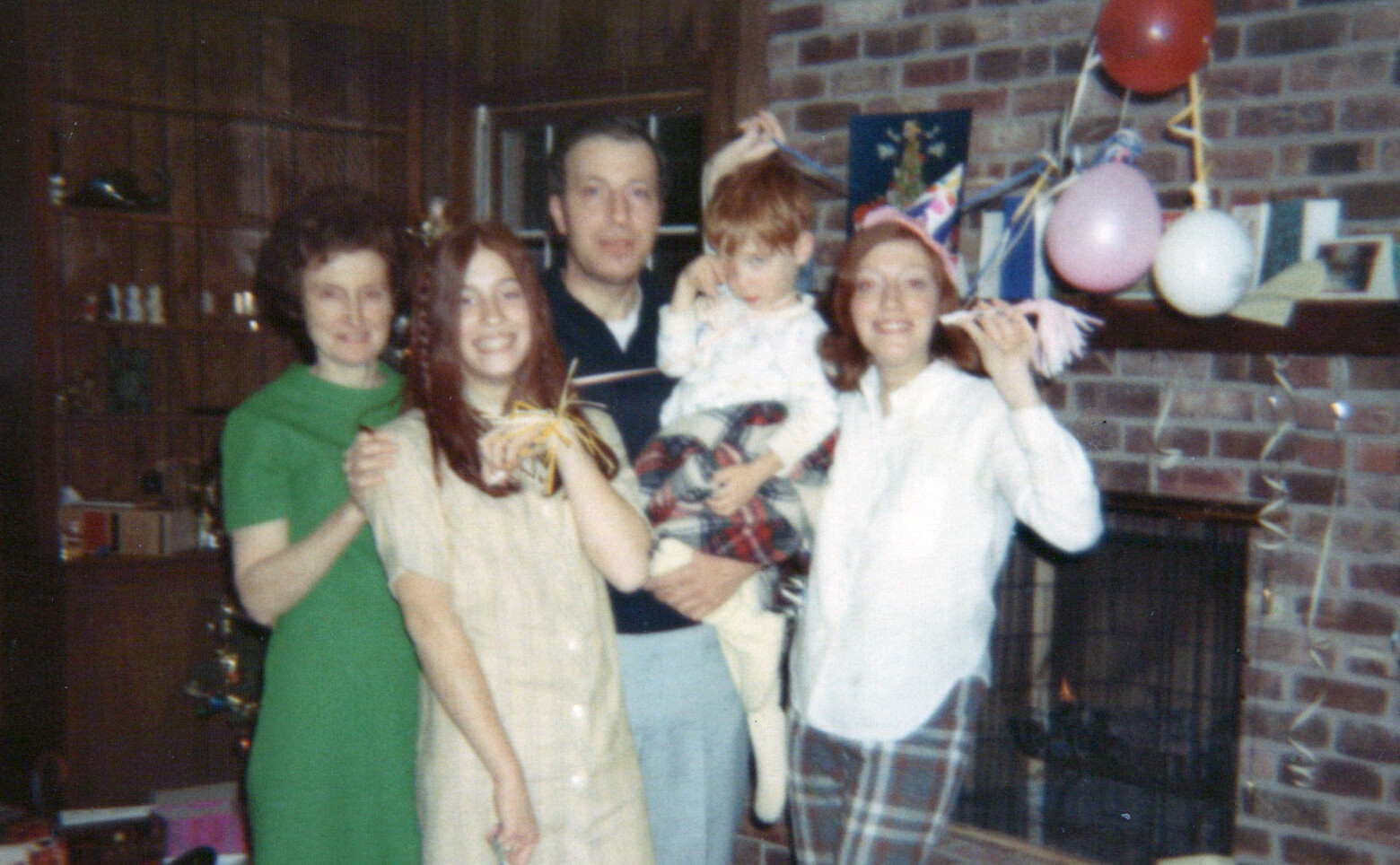 A 1970s family party photo: five adults, a toddler, and a poodle by a brick fireplace with balloons